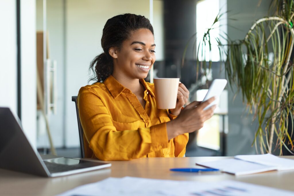 Cheerful african american female manager chatting on cellphone, sitting with cup of coffee in office